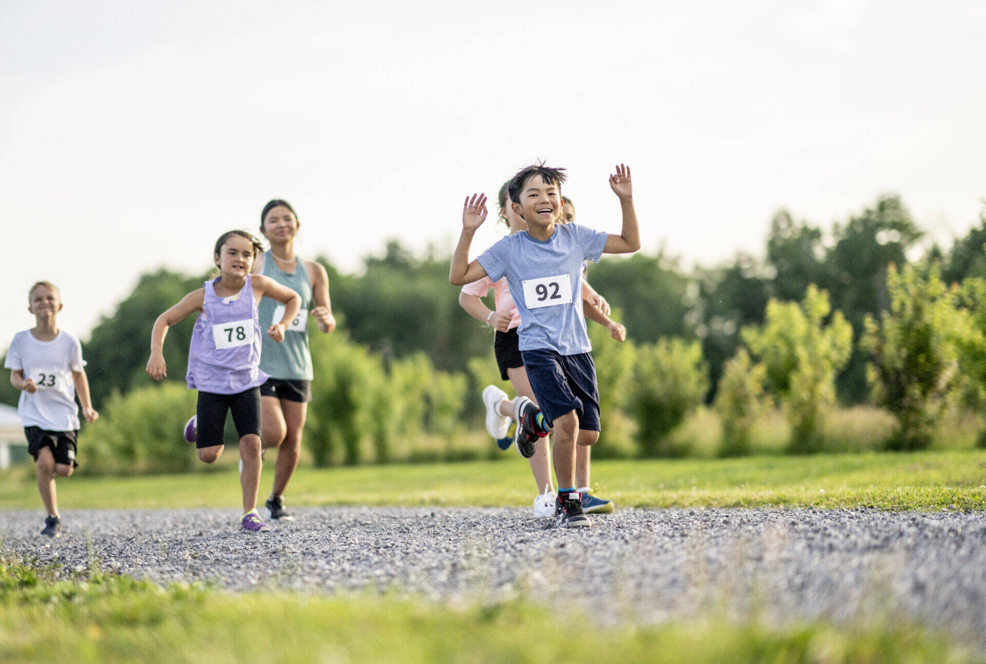 A young boy raises his hands in the air as he crosses the finish line first during a Cross Country race. He is dressed comfortably in athletic wear, have numbered bibs on and his teammates can be seen coming in closely behind him.