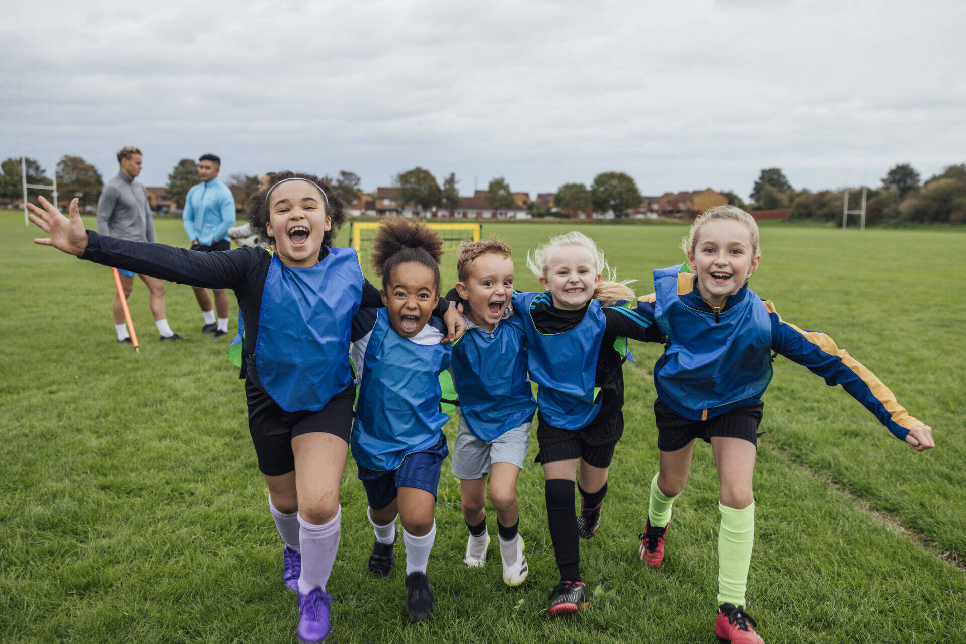 Front view of a small group of boys and girls wearing sports clothing, football boots and a sports bib on a football pitch in the North East of England. They are at football training where they are doing different football training drills. They are running and celebrating with their arms around each other in front of a football goal while smiling and looking at the camera. Videos are available for this scenario.